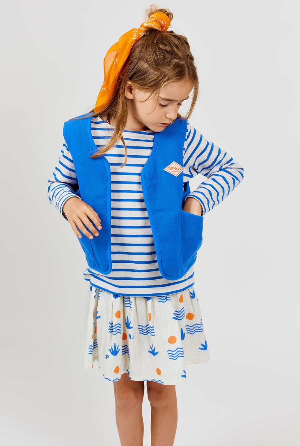 A young girl wears the Arsene & Les Pipelettes Girls' Aloe Skirt featuring a gold elasticated waistband, paired with a striped long-sleeve shirt, a blue vest, and an orange scarf tied in her hair. She looks down with one hand tucked in her vest pocket against a simple background.