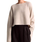 A person is standing against a plain background, wearing an Altuzarra Trip Sweater in an oversized beige cashmere design, paired with black high-waisted trousers.