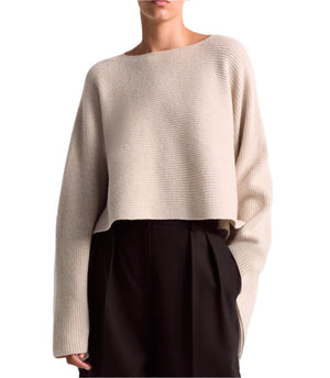 A person is standing against a plain background, wearing an Altuzarra Trip Sweater in an oversized beige cashmere design, paired with black high-waisted trousers.