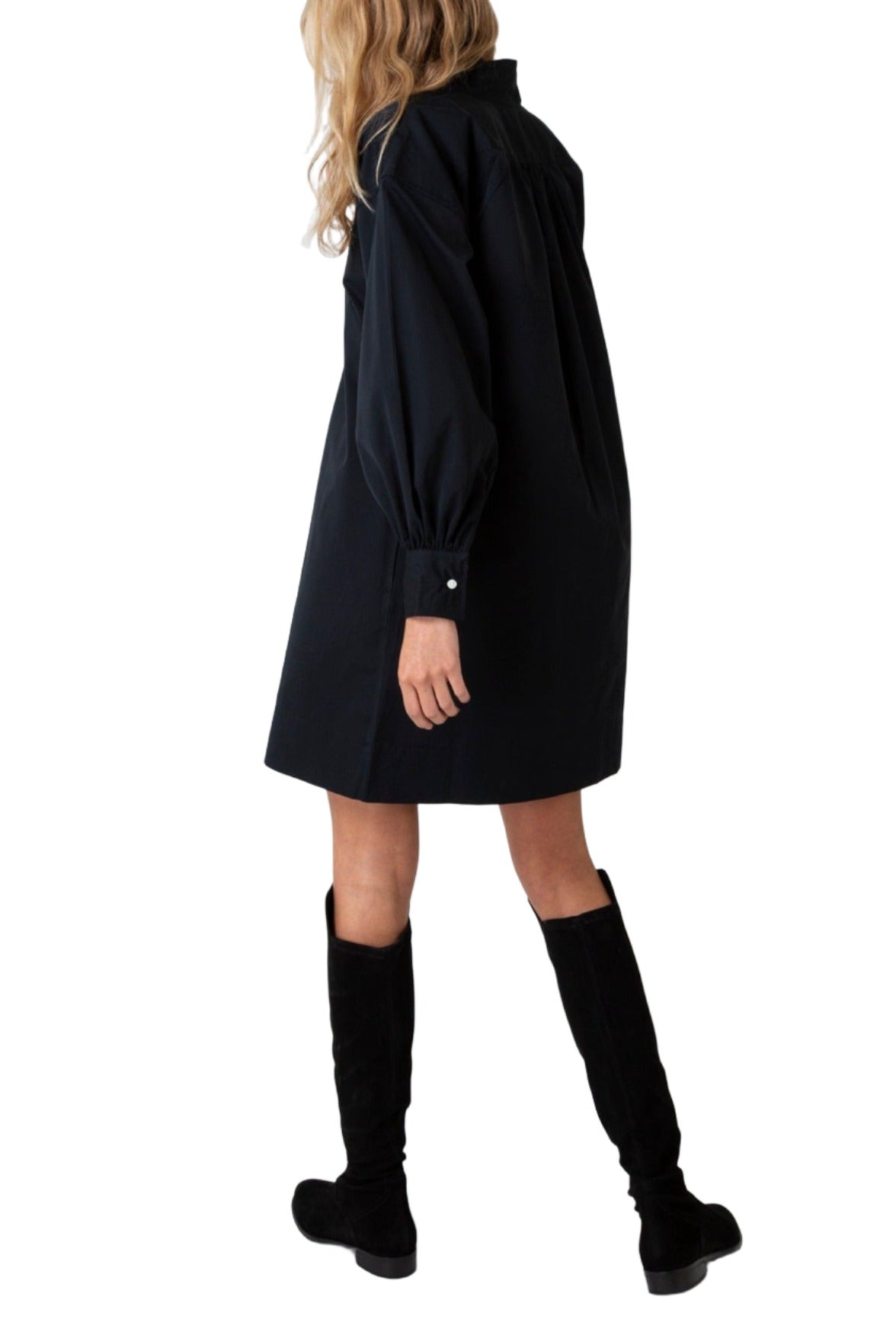 A woman in an Ann Mashburn Anaya Popover dress and knee-high boots, viewed from the side, isolated on a white background.