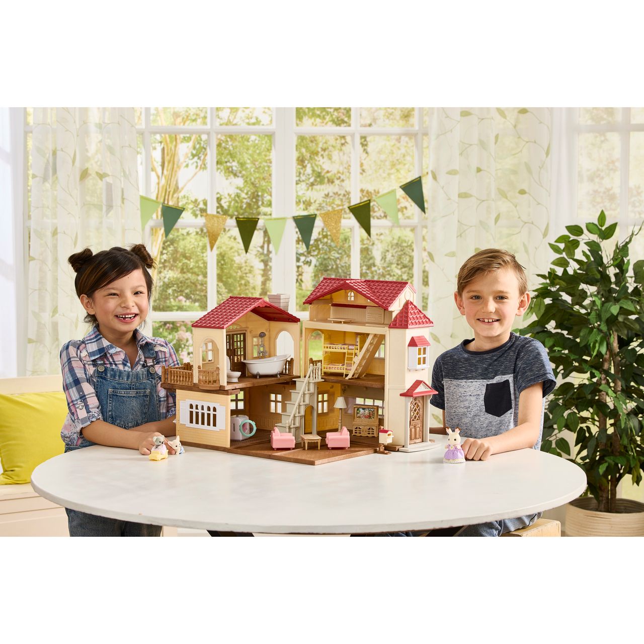 Two children smile as they play with the Calico Critters Red Roof Country Home Gift Set and animal figurines on a round white table in a bright room decorated with green bunting and large windows.