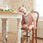 A young girl wears the Kidiwi Ariel Wool Cardigan, sitting on a wooden chair and leaning on a white table in a vintage-style room with patterned floor and floral wallpaper.
