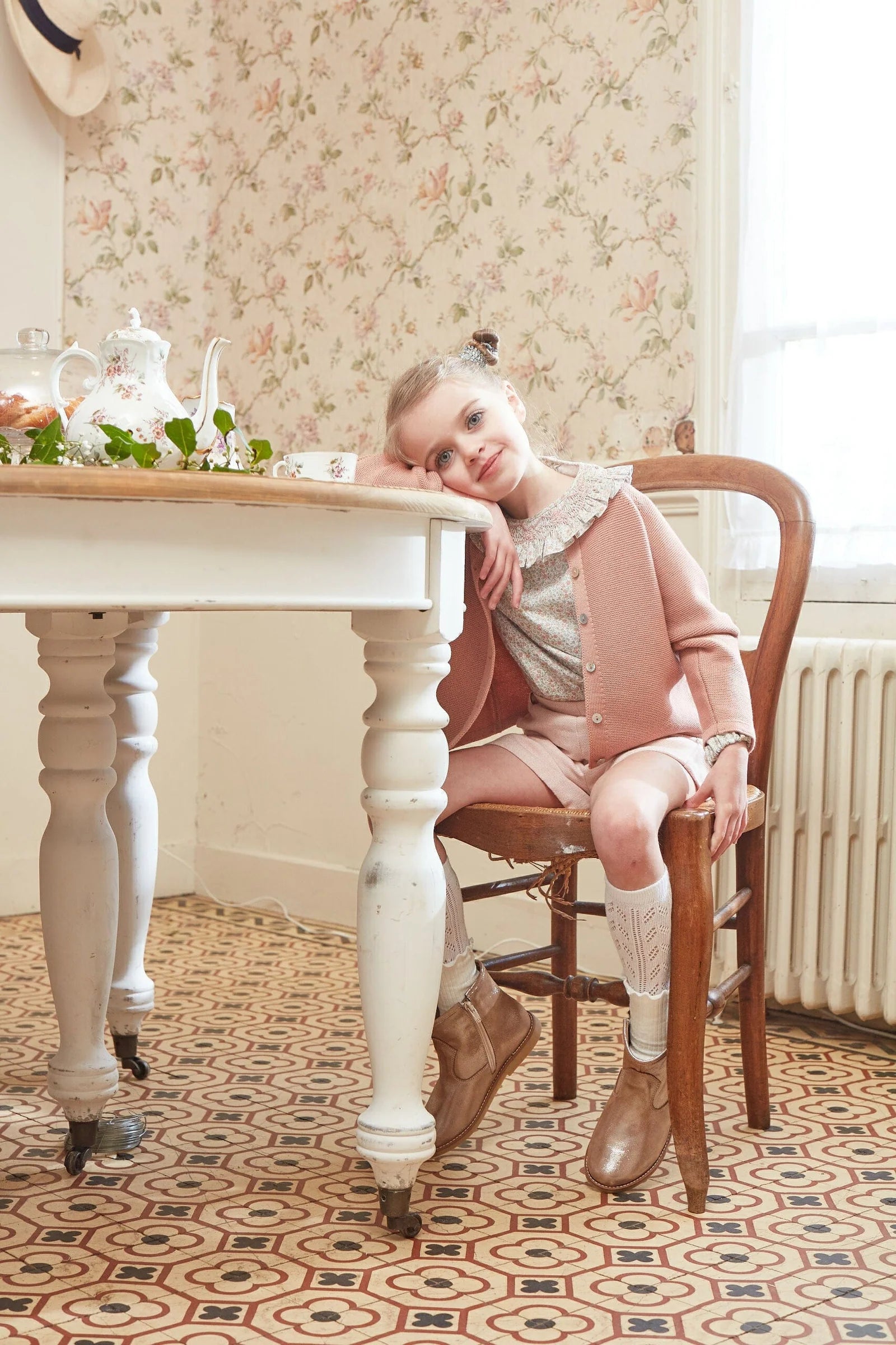 A young girl wears the Kidiwi Ariel Wool Cardigan, sitting on a wooden chair and leaning on a white table in a vintage-style room with patterned floor and floral wallpaper.