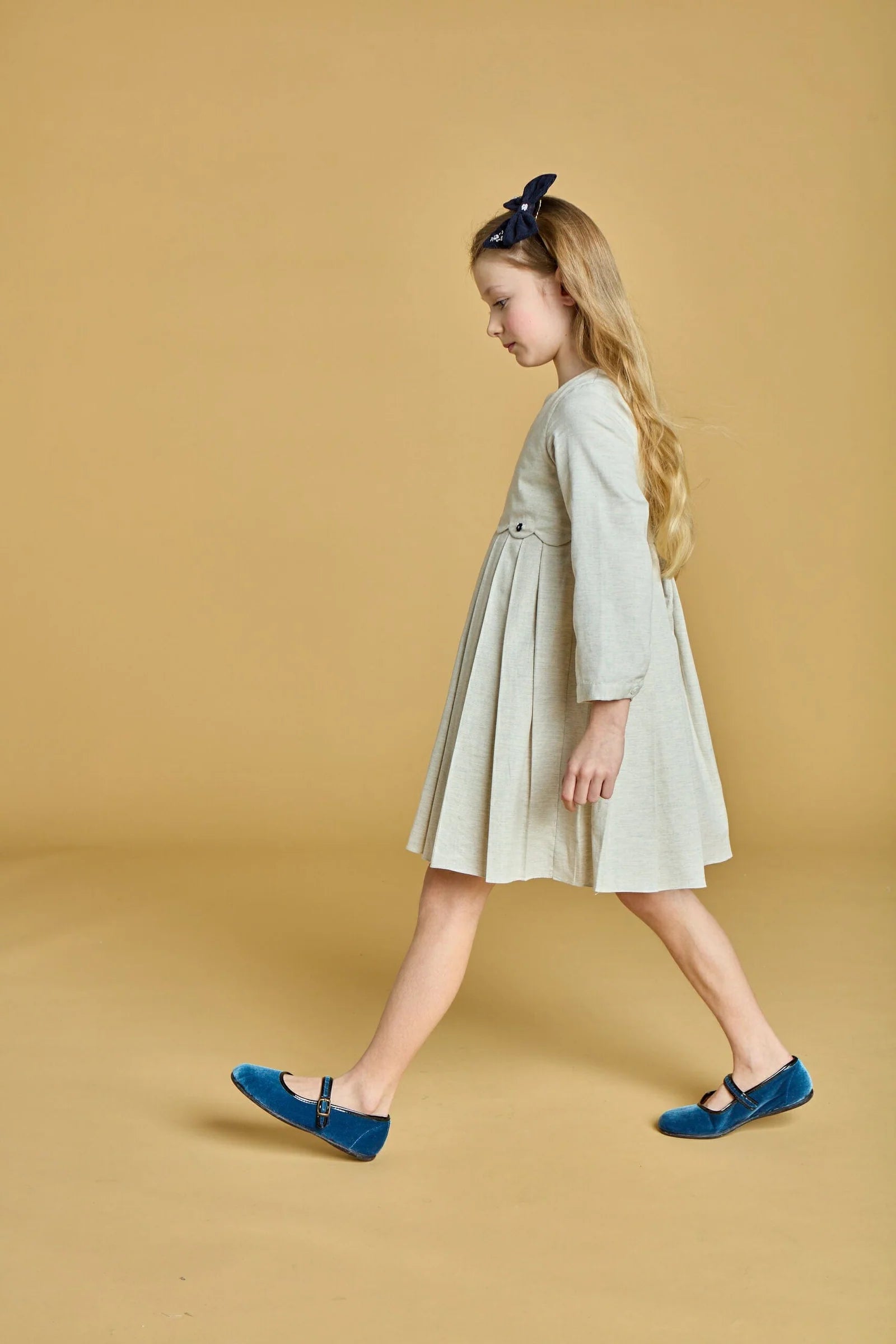 A young girl with long blonde hair, blue shoes, and a dark headband walks against a beige background, wearing the Kidiwi Girls' Ashley Organic Cotton Twill Dress in grey melange.