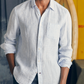 A man in the Faherty Laguna Linen Shirt, a lightweight light blue and white striped style with rolled sleeves, stands indoors near a surfboard.