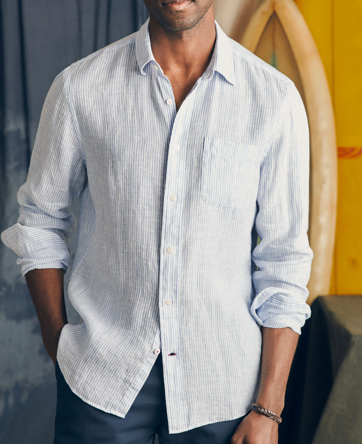 A man in the Faherty Laguna Linen Shirt, a lightweight light blue and white striped style with rolled sleeves, stands indoors near a surfboard.