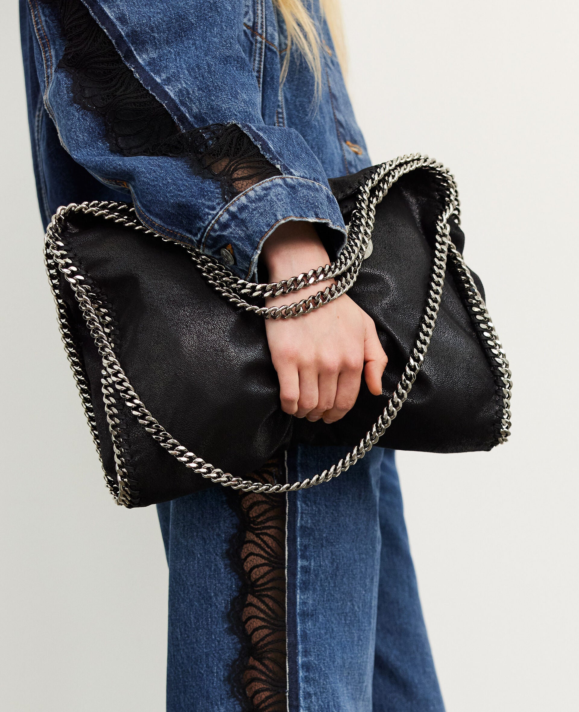 A person in blue denim with black lace details carries the Stella McCartney Falabella Fold Over Tote, a vegan bag made from recycled plastic bottles, featuring bold silver chain handles.