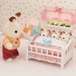 A toy bunny stands beside the Calico Critters Crib with Mobile, where three smaller toy bunnies are tucked under a pink blanket in a pastel nursery setting, creating the perfect baby nursery set for your dollhouse furniture collection.