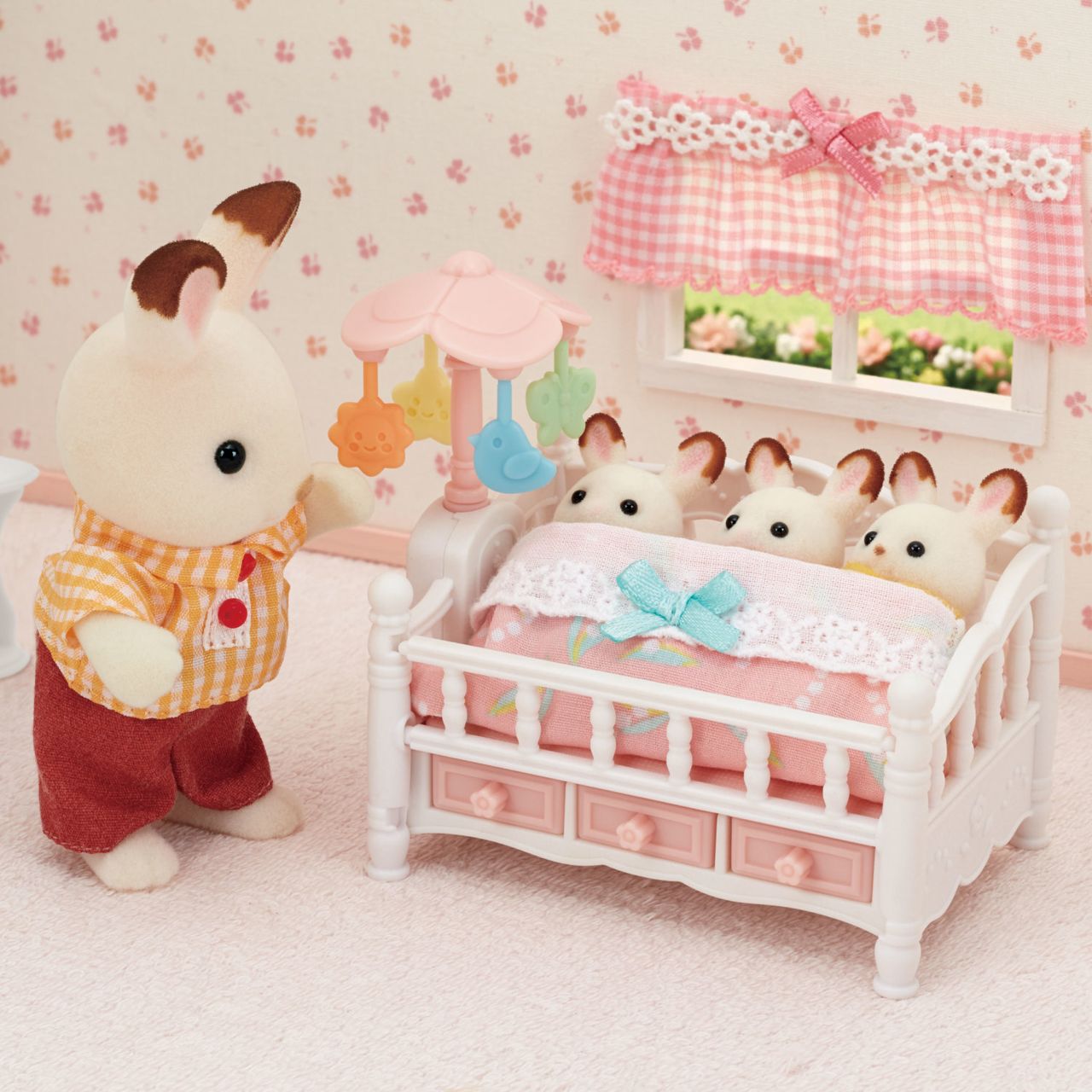 A toy bunny stands beside the Calico Critters Crib with Mobile, where three smaller toy bunnies are tucked under a pink blanket in a pastel nursery setting, creating the perfect baby nursery set for your dollhouse furniture collection.
