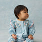A toddler wears the Louise Misha Baby Girls’ Lilette Jumpsuit—crafted from blue cotton linen with an embroidered yoke and ruffled sleeves—paired with floral socks and pink shoes, as she sits on a light blue surface against a matching blue background, looking to the right.