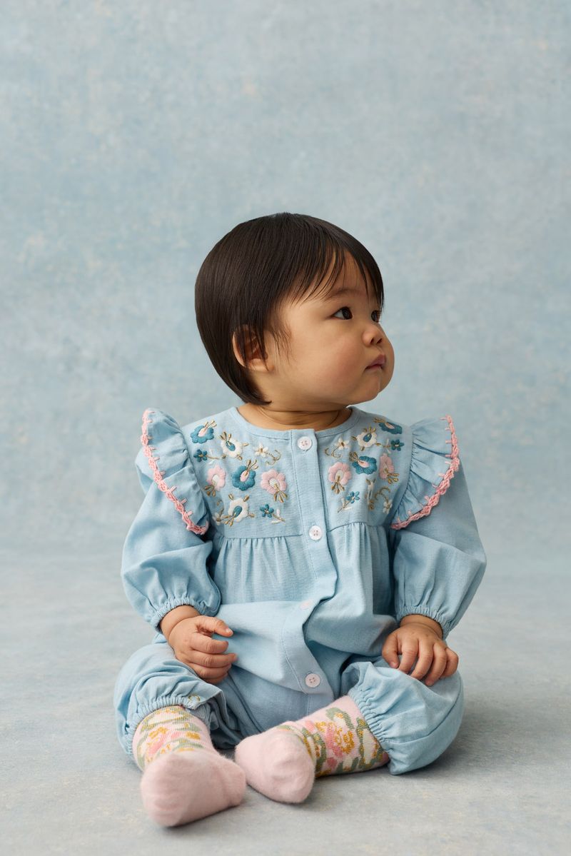A toddler wears the Louise Misha Baby Girls’ Lilette Jumpsuit—crafted from blue cotton linen with an embroidered yoke and ruffled sleeves—paired with floral socks and pink shoes, as she sits on a light blue surface against a matching blue background, looking to the right.