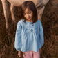 A young girl wears the Louise Misha Girls’ Risa Blouse—crafted from blue cotton and linen with puffy sleeves and floral embroidery—as she stands in tall grass in front of a white horse.