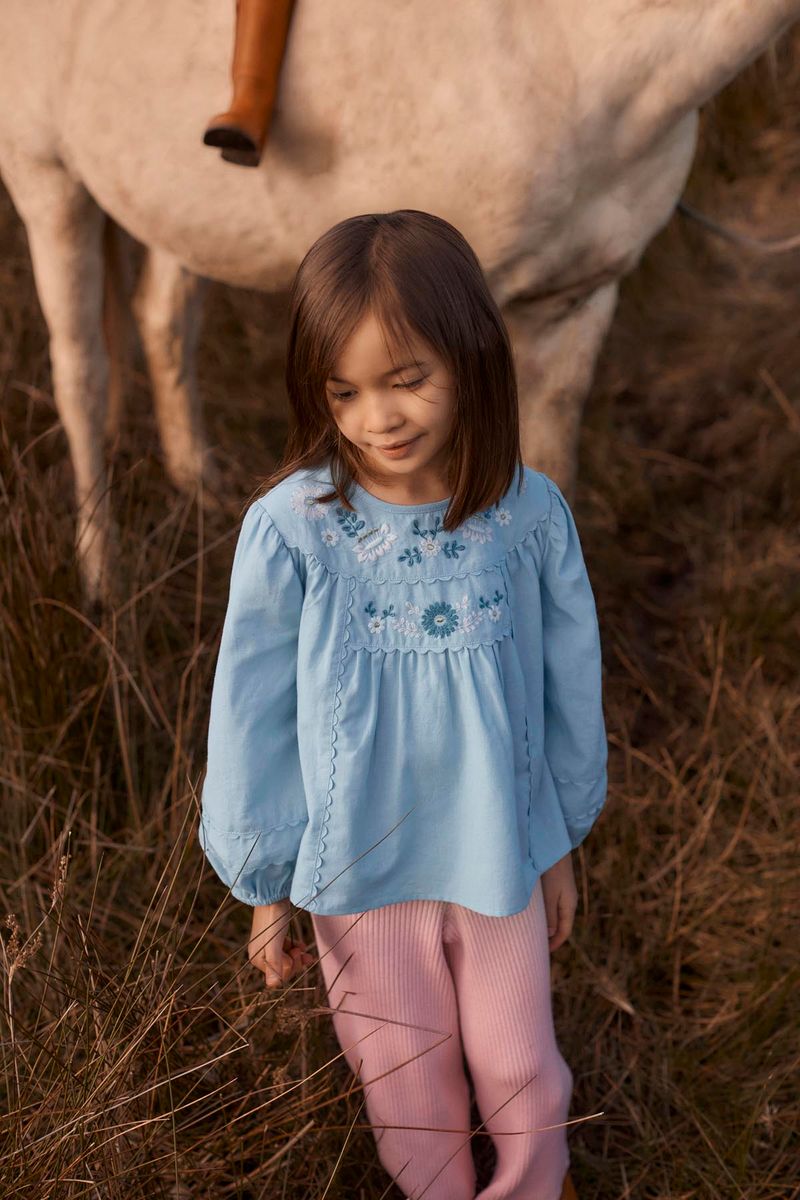 A young girl wears the Louise Misha Girls’ Risa Blouse—crafted from blue cotton and linen with puffy sleeves and floral embroidery—as she stands in tall grass in front of a white horse.