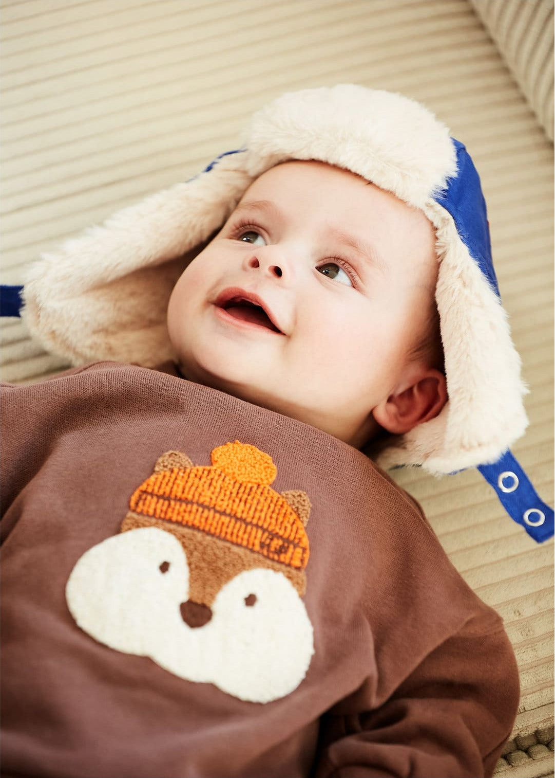 A baby lies on a beige surface wearing Arsene & Les Pipelettes' Baby Boys' Gibson Squirrel Sweatshirt and a blue hat with ear flaps. The comfy animal sweatshirt with an orange knit hat adds extra charm to his adorable look.