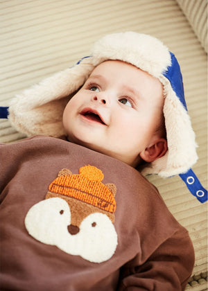 A baby lies on a beige surface wearing Arsene & Les Pipelettes' Baby Boys' Gibson Squirrel Sweatshirt and a blue hat with ear flaps. The comfy animal sweatshirt with an orange knit hat adds extra charm to his adorable look.