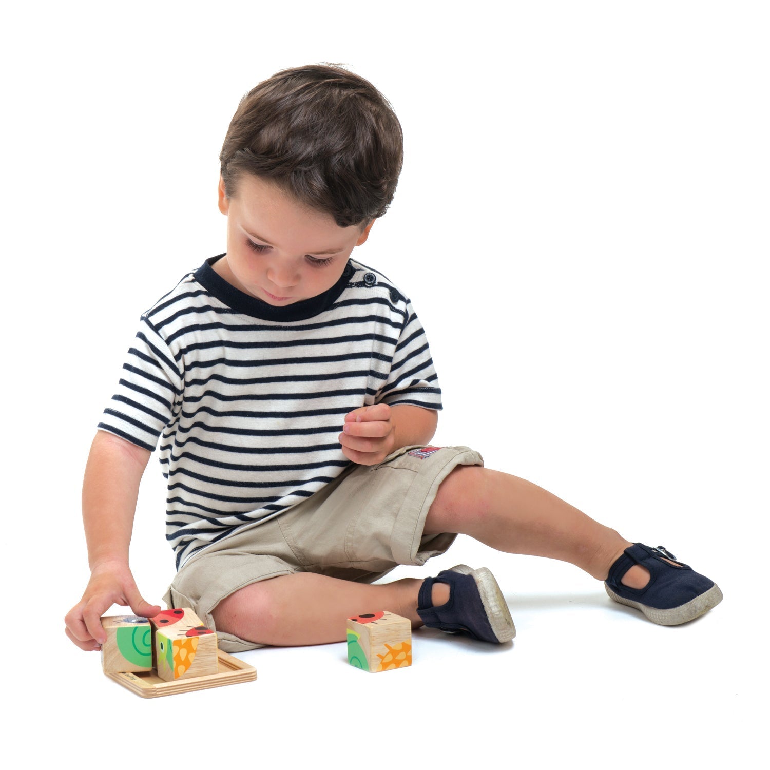 A young child in a striped shirt and khaki shorts sits on the floor, playing with Tenderleaf Baby Blocks from Tender Leaf Toys, a colorful animal block puzzle that enhances hand-eye coordination.