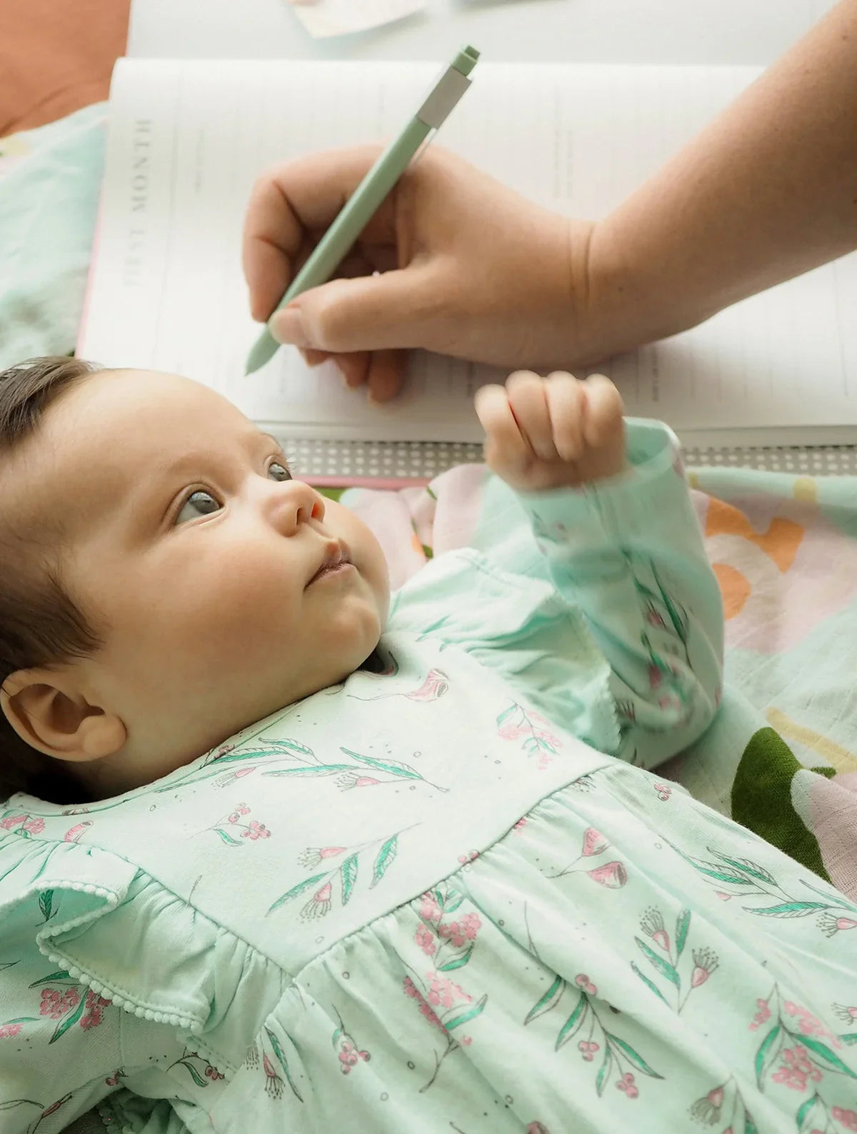 A gender neutral baby is laying on a bed while a woman writes in a notebook, capturing memories for their Write To Me Baby Journal Birth to Five Years Pink.