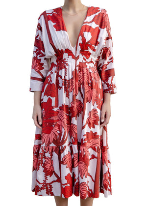 A person wearing an Andres Otalora Batea Dress with a deep V-neckline and long sleeves. The dress, made from soft cotton poplin, features a bold red floral pattern on a white background.