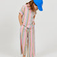A child stands barefoot and looks downward, wearing Arsene & Les Pipelettes Girls' Bayadere Stripe Pants crafted from soft cotton voile with colorful stripes, paired with a blue cap and a short-sleeve button-up shirt.