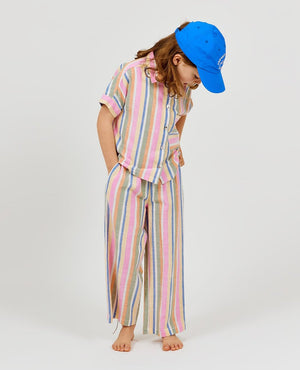 A child stands barefoot and looks downward, wearing Arsene & Les Pipelettes Girls' Bayadere Stripe Pants crafted from soft cotton voile with colorful stripes, paired with a blue cap and a short-sleeve button-up shirt.