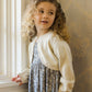 A young child with curly hair, gazing out a window, wears a floral dress topped with the Noralee Girls' Bolero Sweater.