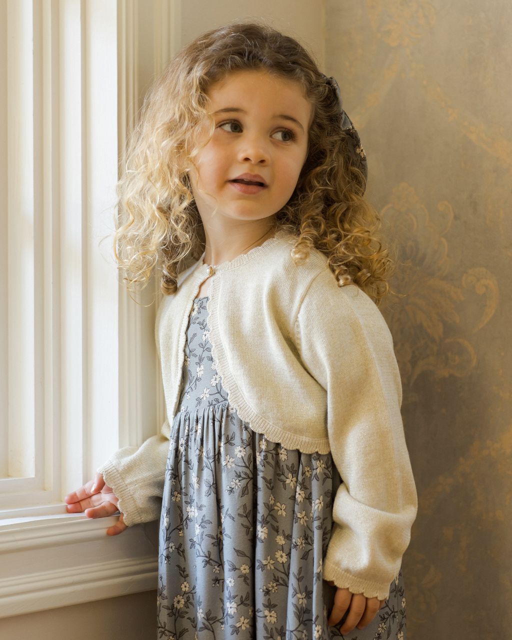 A young child with curly hair, gazing out a window, wears a floral dress topped with the Noralee Girls' Bolero Sweater.
