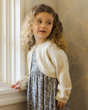 A young child with curly hair, gazing out a window, wears a floral dress topped with the Noralee Girls' Bolero Sweater.