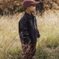 A young boy wears the Rylee & Cru Baby Boys’ and Boys’ Chore Coat—a classic layering piece with chest patch pockets—paired with striped pants and a maroon cap, standing in tall grass outdoors with trees behind him.