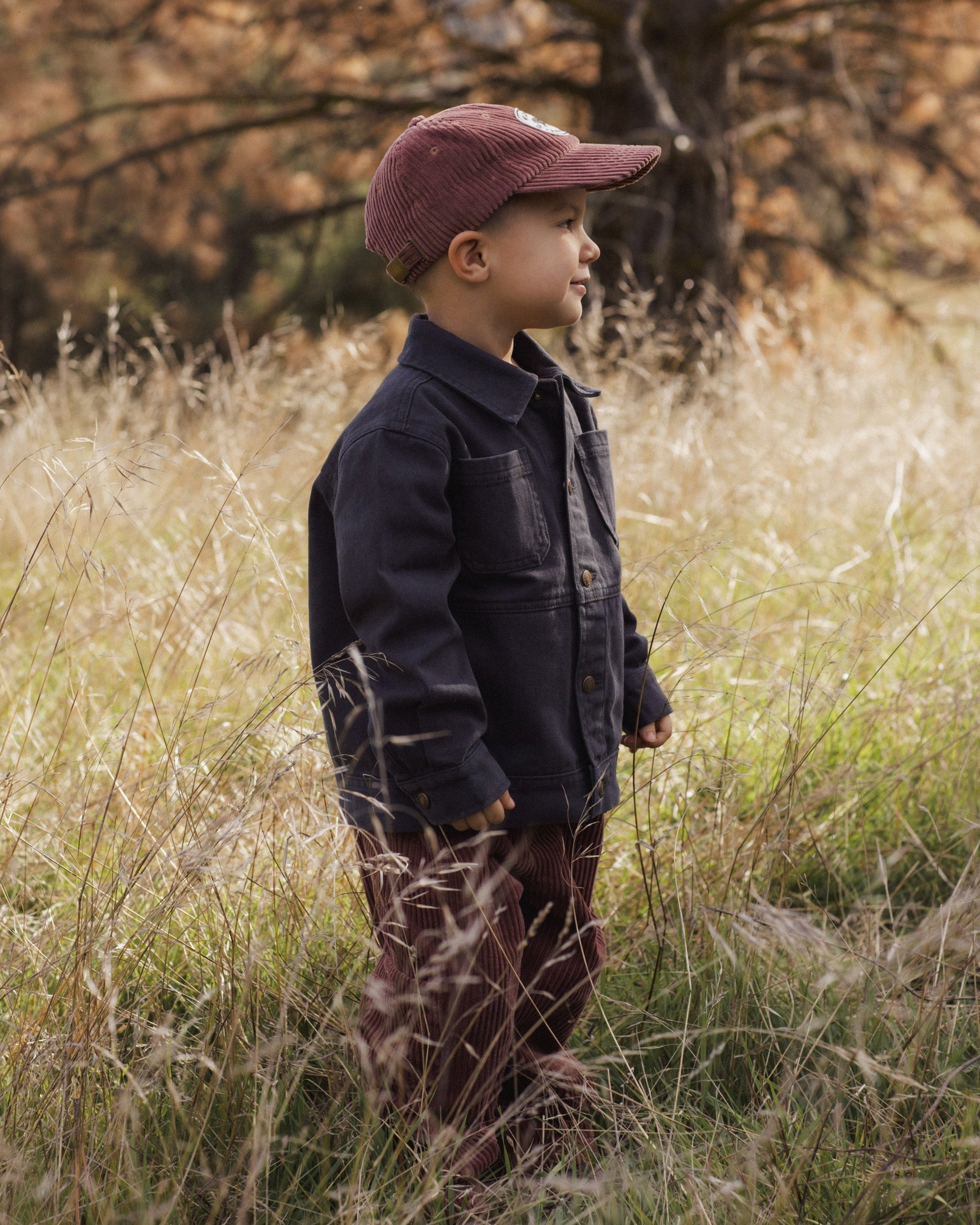 A young boy wears the Rylee & Cru Baby Boys’ and Boys’ Chore Coat—a classic layering piece with chest patch pockets—paired with striped pants and a maroon cap, standing in tall grass outdoors with trees behind him.
