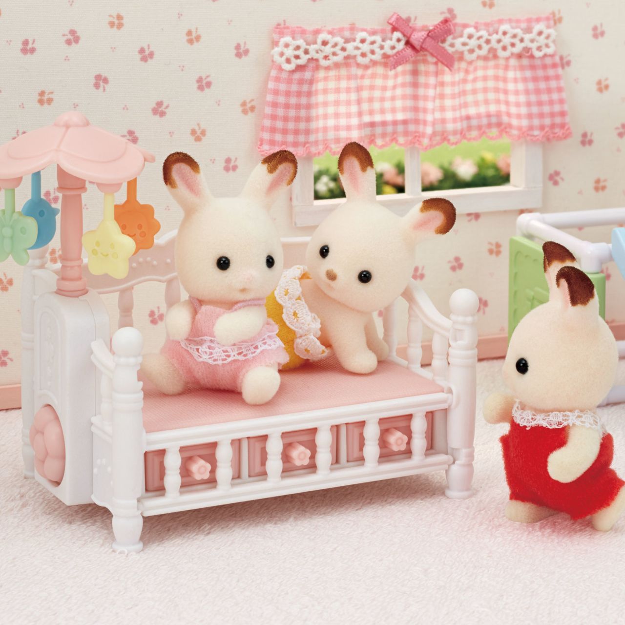 Three Calico Critters plush bunny toys sit and stand around the Crib with Mobile, which features a white frame and a pink mattress, in a pastel-themed dollhouse nursery—an adorable baby nursery set perfect for enhancing any collection of dollhouse furniture.