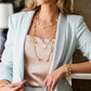 A woman in a light blue blazer and beige top wears the Juliska Capucine De Wulf Berry Clover Double Sided Necklace, layered with double-sided clovers, paired with a gold bracelet featuring white accents.
