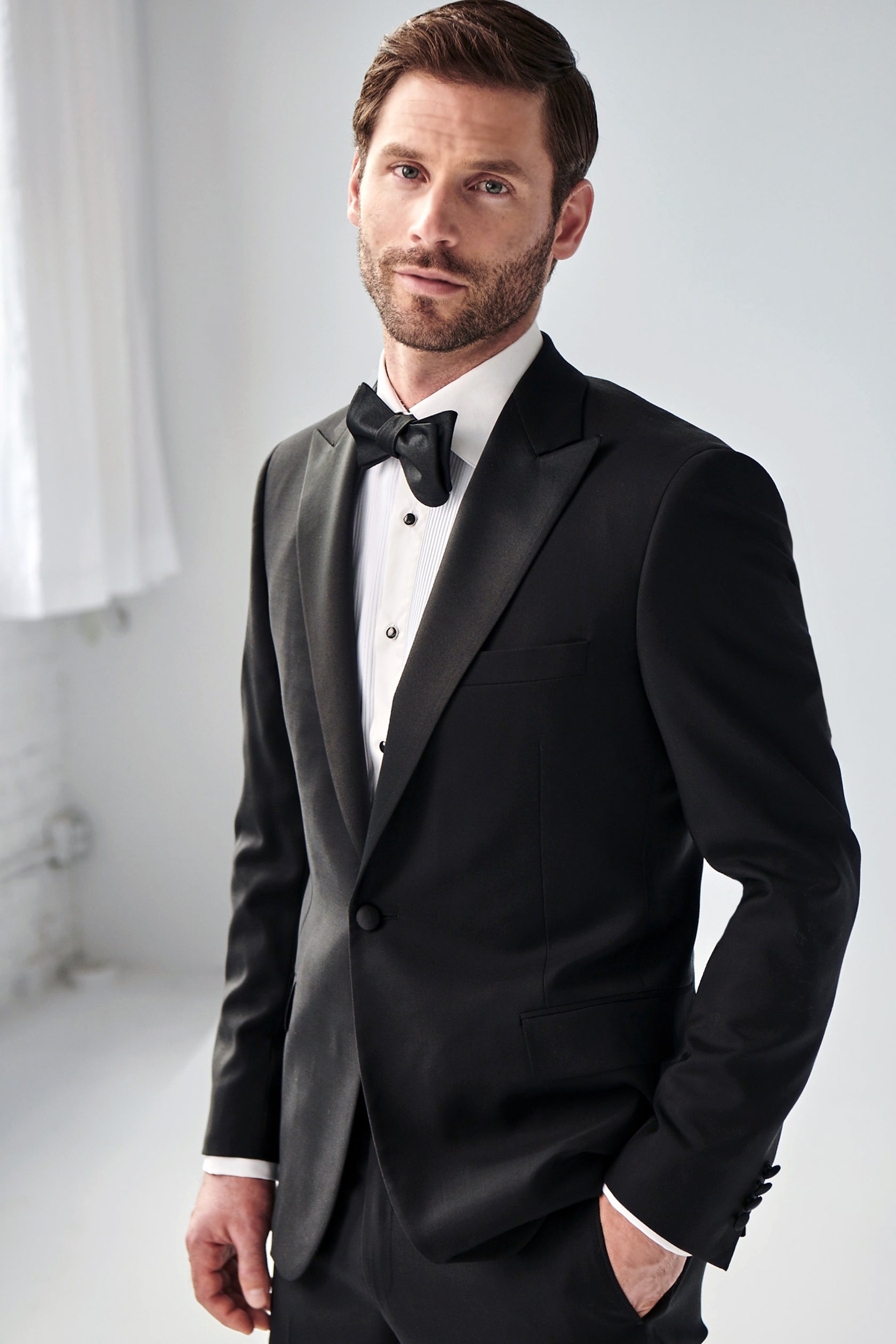 A man in a Samuelsohn Bennet Contemporary Fit Tuxedo with a bow tie posing confidently in a bright room.