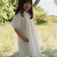A young girl stands outdoors on grass, smiling and holding a balloon string while wearing the Early Sunday Girls’ Beth Dress. Trees and a blue sky are visible in the background.
