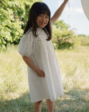 A young girl stands outdoors on grass, smiling and holding a balloon string while wearing the Early Sunday Girls’ Beth Dress. Trees and a blue sky are visible in the background.