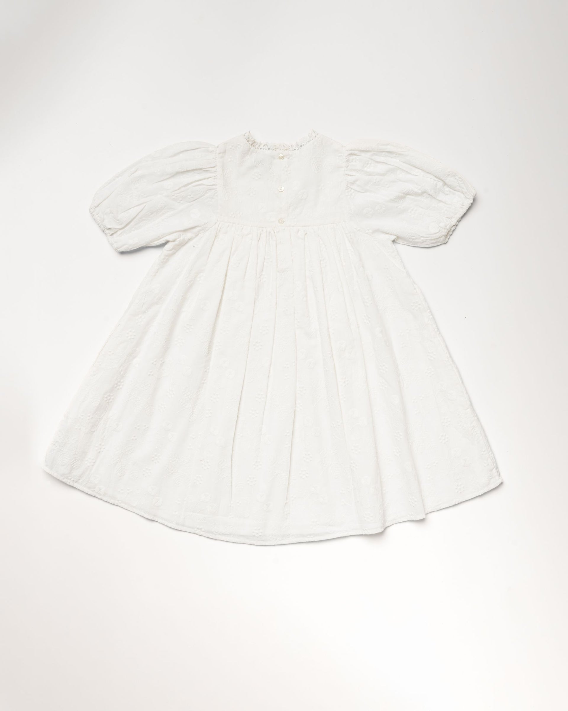 Early Sunday Girls’ Beth Dress in ivory, featuring short sleeves, a high collar, a gathered waist, and subtle embroidered cotton details, laid flat on a plain background.