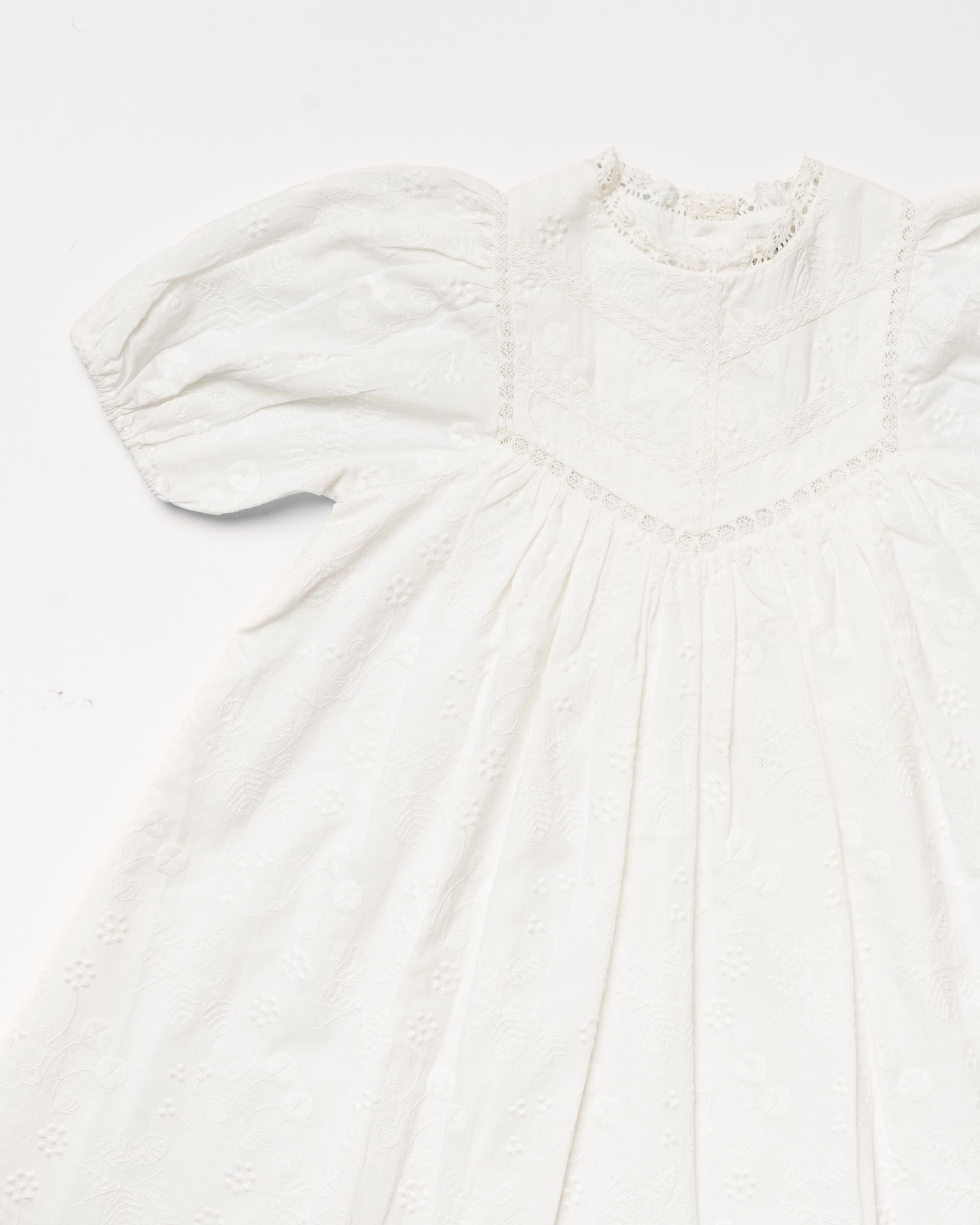 The Early Sunday Girls’ Beth Dress in ivory cotton features a vintage-inspired design with puffed sleeves, a high neckline, lace trim, and delicate floral embroidery. Shown laid flat on a plain white background.