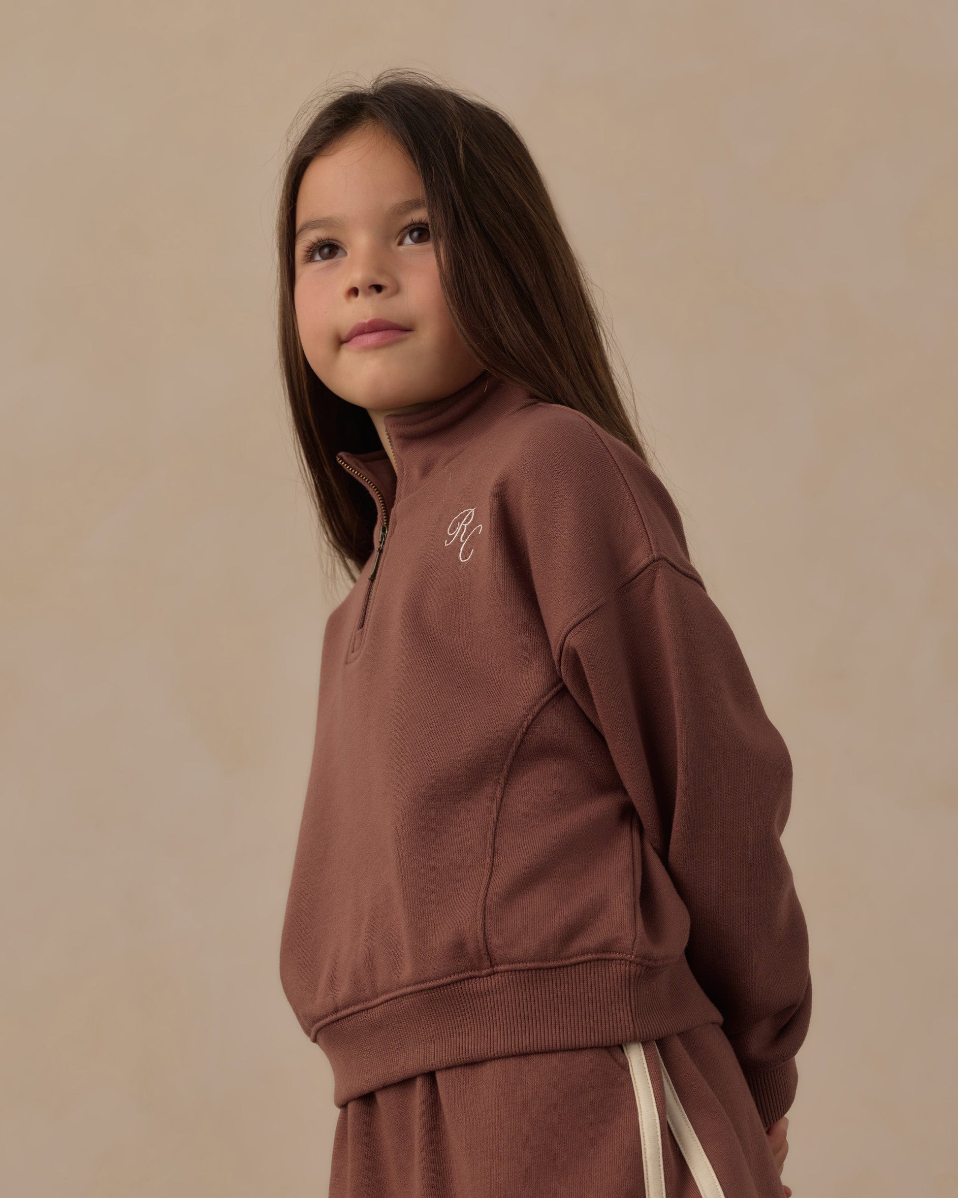 A young girl with long brown hair wears the Rylee & Cru Girls’ Half Zip Sweatshirt in a sporty mauve French terry, paired with matching pants, while standing against a neutral beige background.