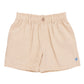 The Boys’ Jackson Short by Pink Chicken features beige cotton canvas, an elastic waistband, button closure, belt loops, and a small blue embroidered logo near the left hem.