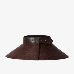 The Janessa Leone Birdie Visor is a wide-brimmed brown raffia straw hat featuring a dark leather band with metal studs around the crown, shown against a plain white background.