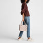 A person wearing a maroon top, blue jeans, and white sneakers stands holding a light pink Bitty Bogg® Bag by Bogg Bags—a compact tote featuring signature cutout holes and designed to be easy to clean as a washable bag.