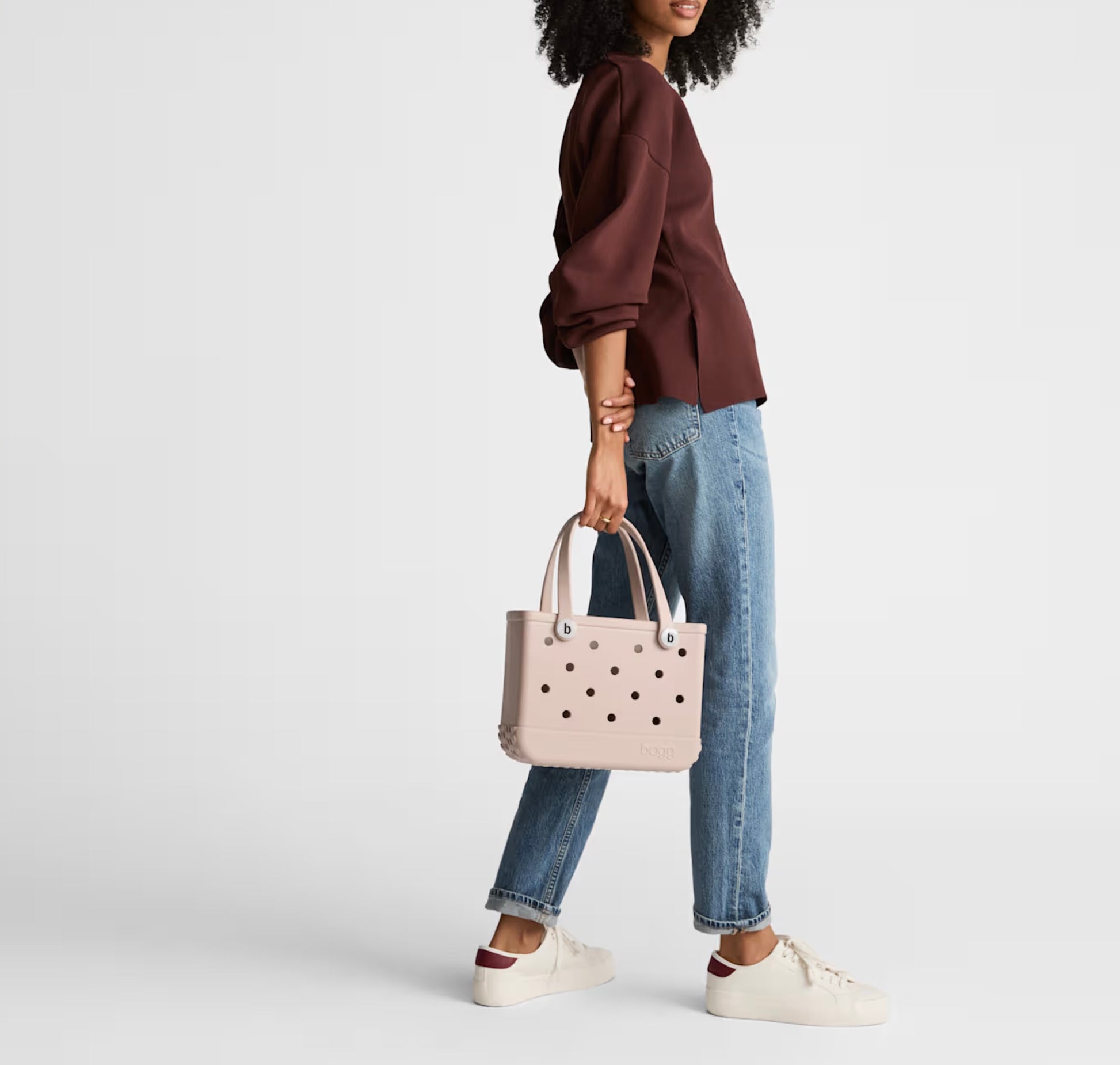 A person wearing a maroon top, blue jeans, and white sneakers stands holding a light pink Bitty Bogg® Bag by Bogg Bags—a compact tote featuring signature cutout holes and designed to be easy to clean as a washable bag.