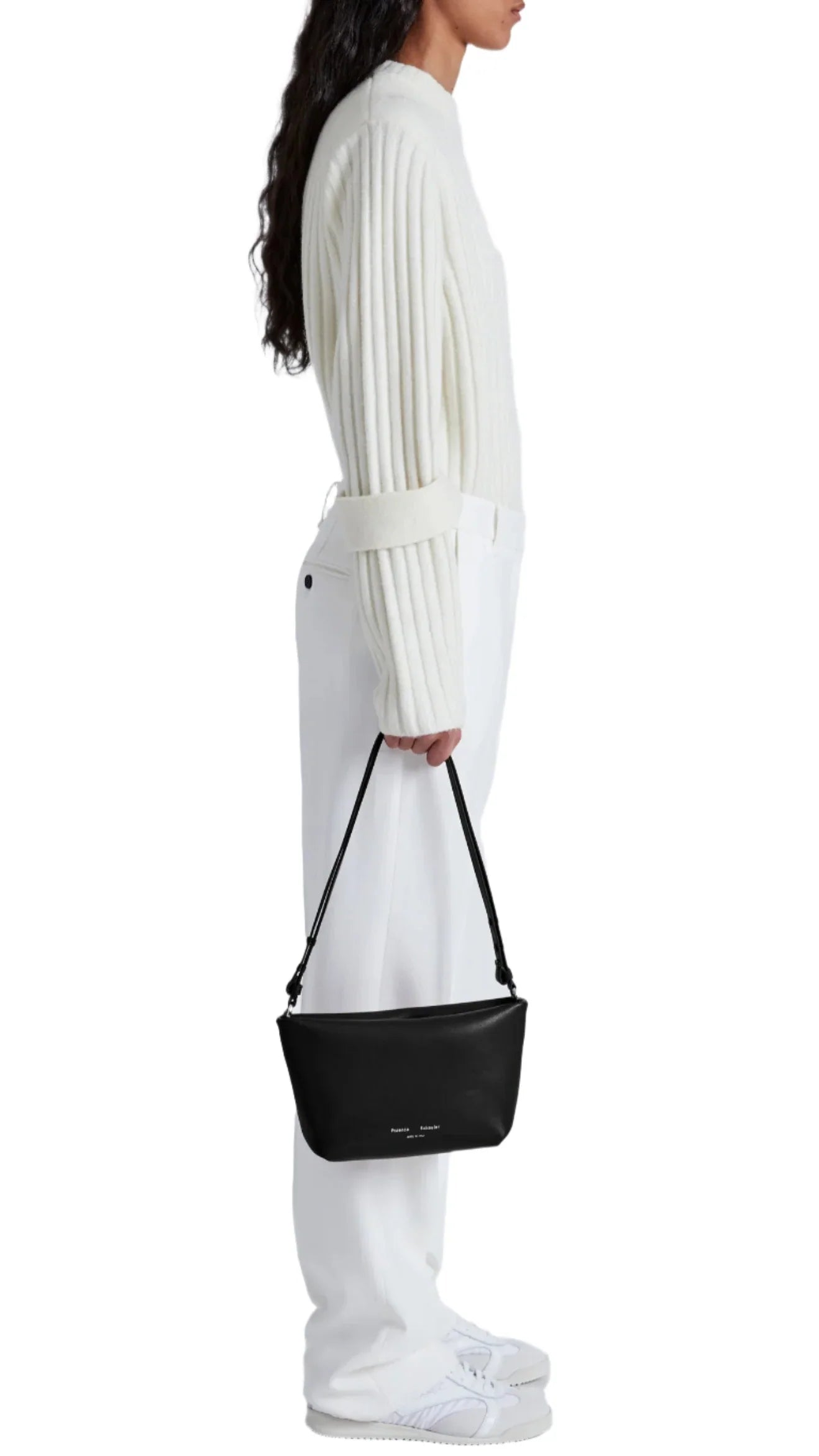 A person stands in profile, dressed in a white ribbed sweater, white pants, and white shoes, holding a small black Proenza Schouler Bond Smooth Nappa Bag with supple leather and shoulder to crossbody function down by their side.