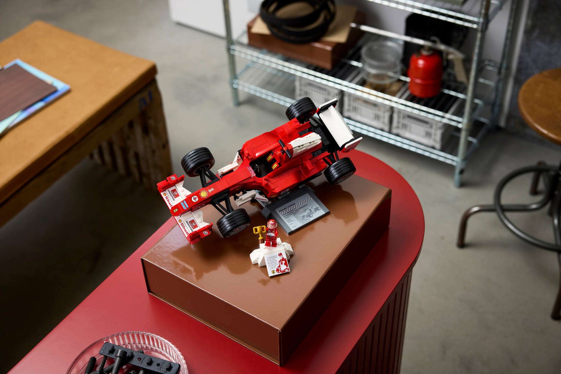 The LEGO® Icons Ferrari F2004 & Michael Schumacher set by Legos - Toyhouse features a model car and minifigure displayed on a brown box in a modern room with shelves and furniture.