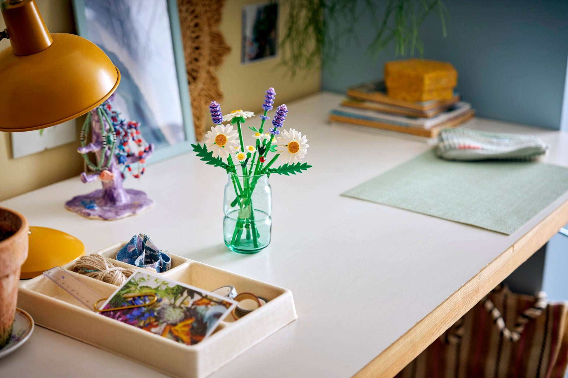 A tidy desk with a yellow lamp, a jar of Legos - Toyhouse LEGO® Botanicals Daisies artificial flowers, a white organizer tray, stacked books, and a folded cloth on a green mat.