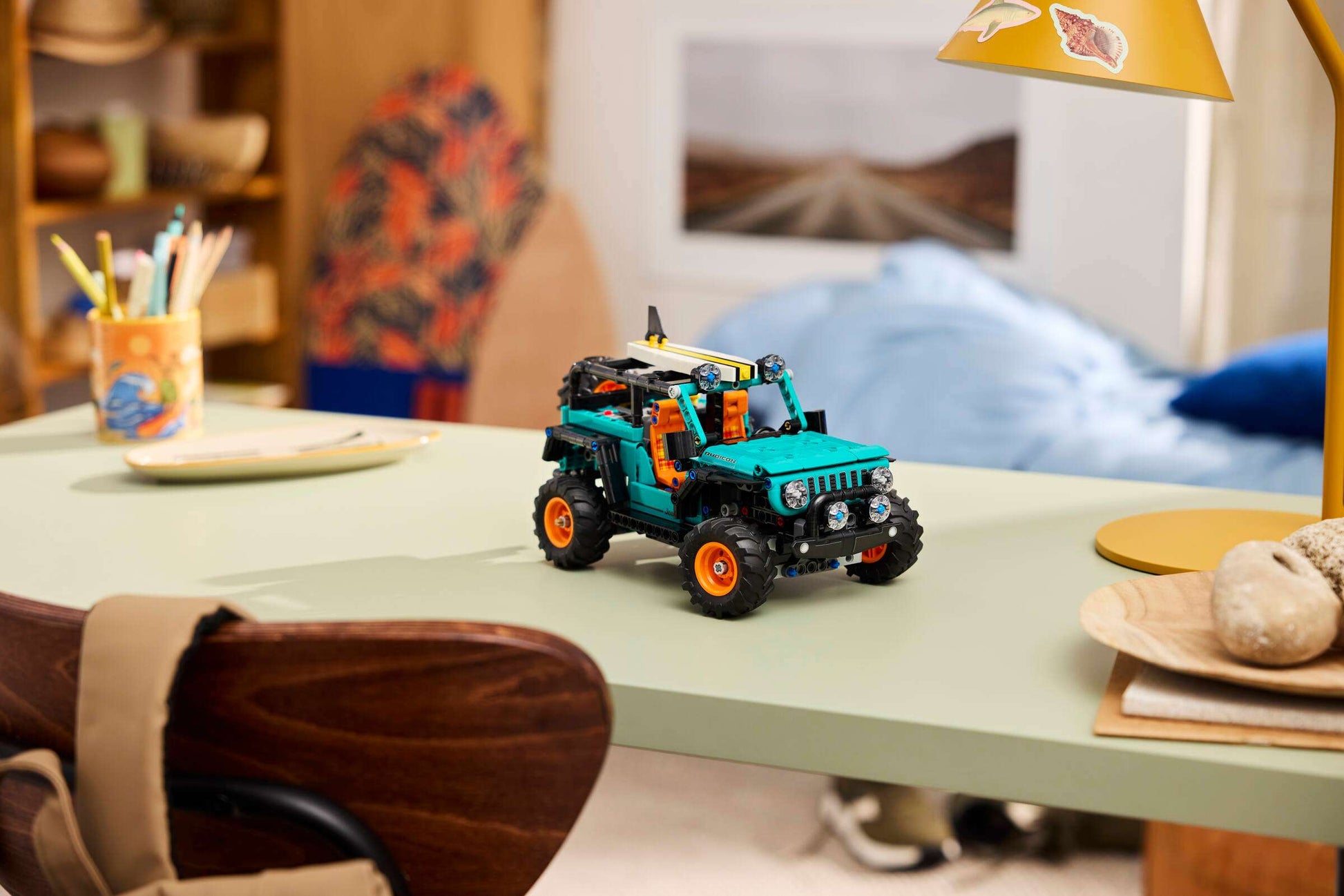 A LEGO® Technic™ Jeep® Wrangler Rubicon SUV by Legos - Toyhouse, with teal and black colors and orange wheels, is displayed on a light green desk in a cozy, well-lit room.