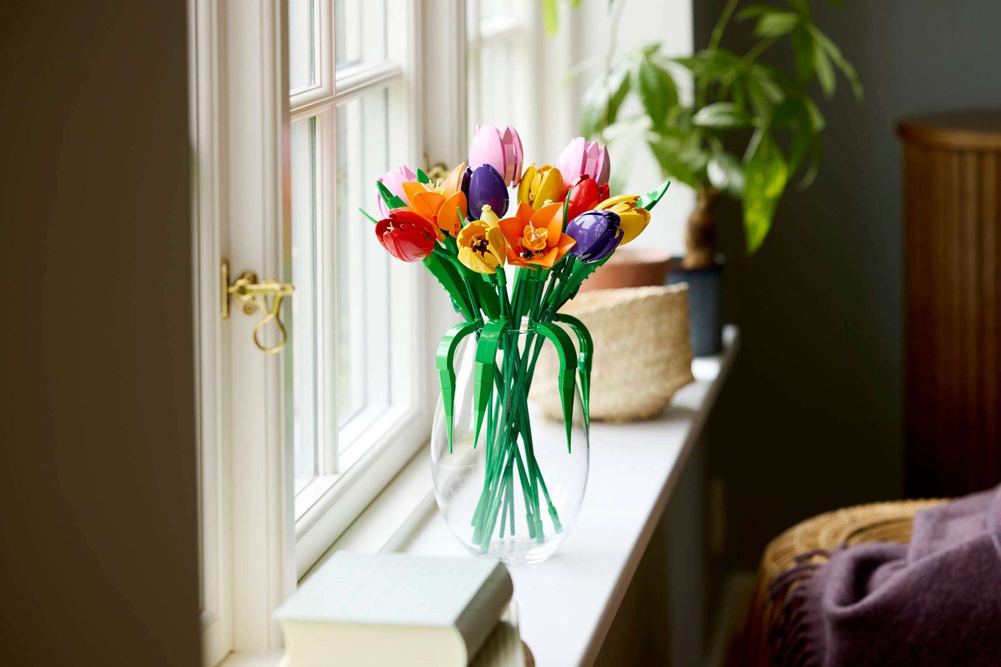 The LEGO® Botanicals Tulip Bouquet by Legos - Toyhouse sits in a clear vase on a windowsill beside a plant, bringing playful floral decor and vibrant color to the sunlit room.