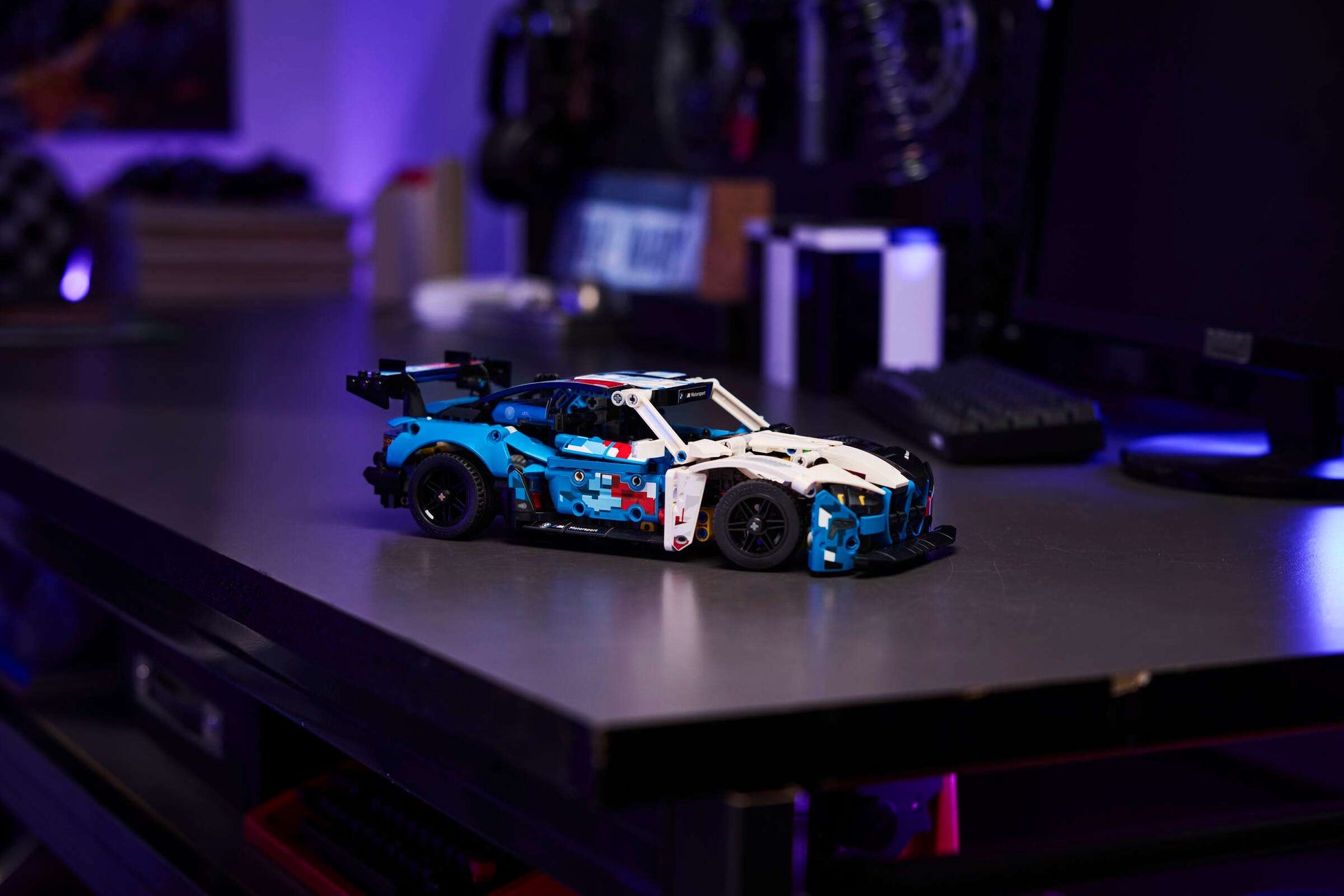 A LEGO® Technic™ BMW M4 GT3 EVO Race Car kit from Legos - Toyhouse rests on a metal desk in a dim room with electronics and computers—a great STEM building toy for fans.