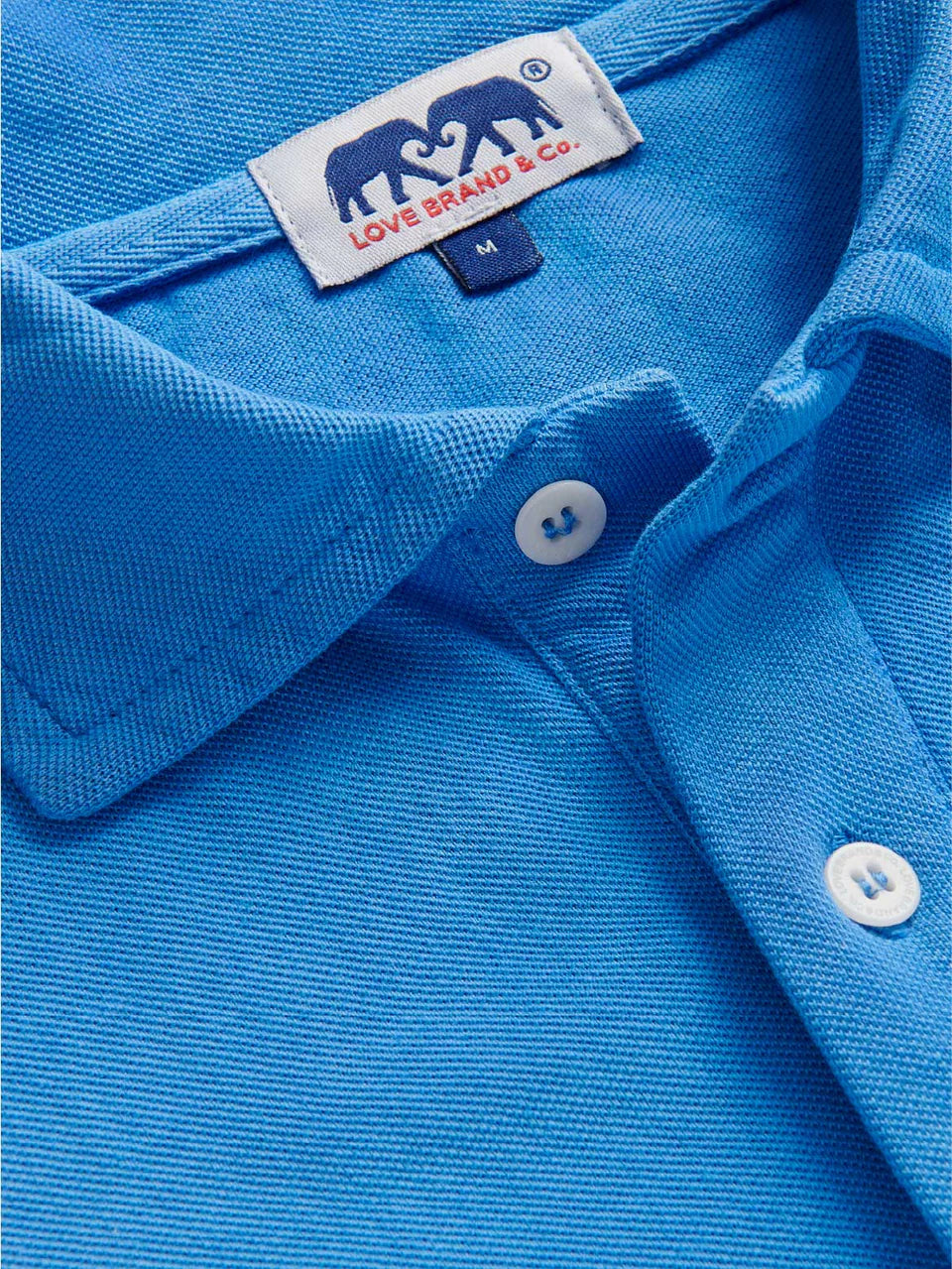 Close-up of the Pensacola Polo Shirt by Love Brand—a blue organic cotton polo with white buttons and a label that features two elephants. A smart-casual essential.