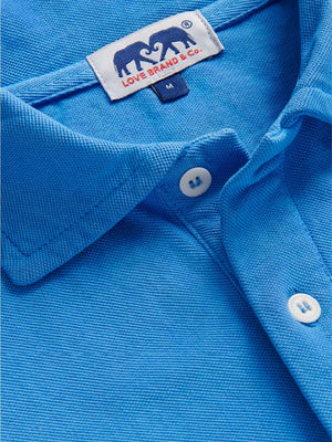 Close-up of the Pensacola Polo Shirt by Love Brand—a blue organic cotton polo with white buttons and a label that features two elephants. A smart-casual essential.