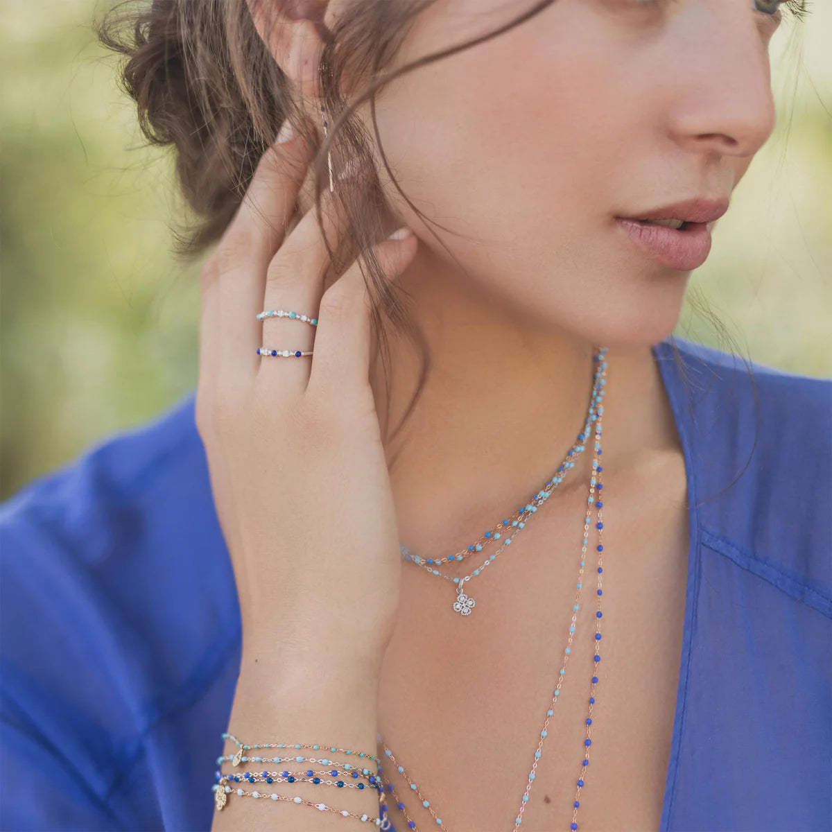 A woman wearing layered blue and silver jewelry, including the Gigi Clozeau Classic Gigi Bracelet 6.7", with necklaces and rings, paired with a blue top—ideal inspiration for everyday jewelry styling.